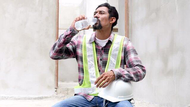 Hopeless middle-aged Indian worker stressed sweat tired thirsty drinking water sitting on cement floor at construction site worried about financial crisis or termination of employment unhealthy man