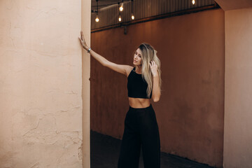 Beautiful blonde model woman, fashionable in a black suit, poses against the walls of a building