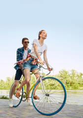 Fototapeta premium Vertical portrait of active young couple riding one bicycle on city street. Lovely, nice riders enjoying themselves. Romantic leisure activity.