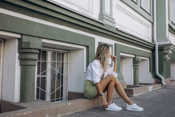 Beautiful blonde model, fashionable in a dress and jacket, poses against the walls of a building