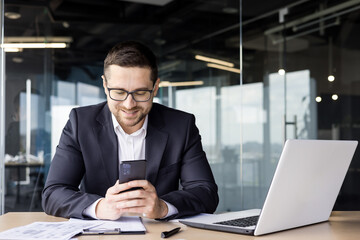 A young male office worker sits at a desk and uses the phone, smilingly typing messages, reading news, playing online games