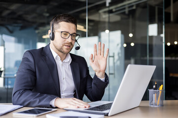 A young man works in the office and wears a headset, sits at a table in front of a laptop, conducts consultations, support service, online business meeting. Says hello and waves to the camera