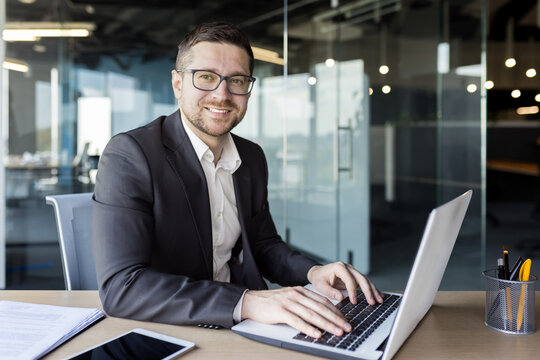 Portrait of a young man sitting at a desk in an office center in a suit and working on a laptop. Looking and smiling at the camera