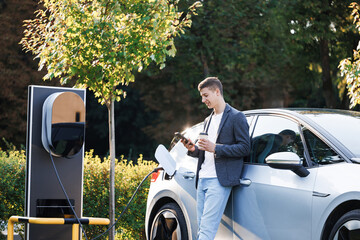 Caucasian businessman standing near an electric car that is charging and making time adjustments on...