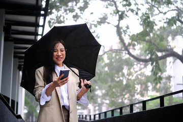 Millennial businesswoman with umbrella using smartphone and walking down city street during rain