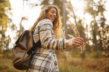 Hand of a stylish tourist with a backpack holds to the soft golden pampas grass in the autumn season. Beautiful woman enjoys studying nature in the autumn forest. Adventure, vacation concept.