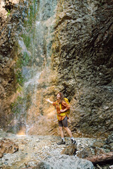 Obraz premium Smiling female traveler with a yellow backpack, dressed in hiking clothes, enjoys a waterfall near a mountain river. Travel, trekking. Nature concept.