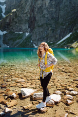 A smiling female traveler with a yellow hiking backpack against the backdrop of a turquoise lake among the mountains. Active lifestyle. Hiking, adventure concept.