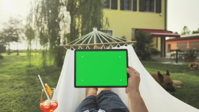 pov of man holding tablet green screen on the hammock in the home garden,point of view of male relaxing in the backyard watching content on chroma key display