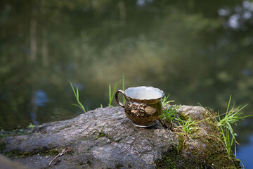 Glass vintage cup in gold color with patterns, stands on a stone. against the backdrop of a picturesque river. Image for your creativity, design, illustrations.