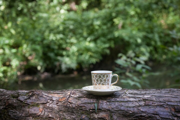 A cup of coffee with golden patterns, in a small saucer, stands on a log, against the backdrop of a picturesque river in the morning. Image for your design or creative illustrations.