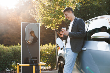 Caucasian businessman using smart phone and waiting power supply connect to electric vehicles for...