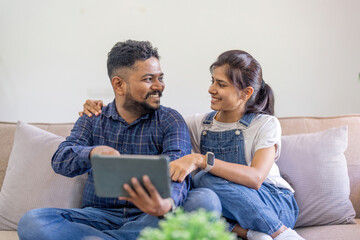 Fototapeta premium Beautiful indian spouses rest on sofa at home, using digital tablet explore the world of social media and shopping online together