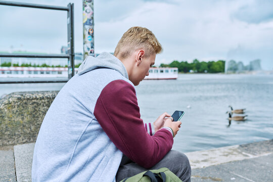 Serious guy college student using smart phone sitting on city river embankment