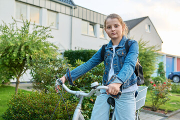 Schoolgirl child 10, 11 years old with backpack on bicycle on street near house