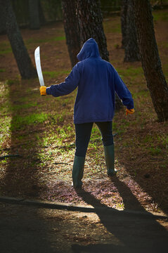 Terrifying Halloween Scene: Hooded Figure in Gardener's Gloves and Rubber Boots Wielding Machete, Haunting the Spooky Forest