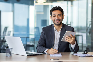 Portrait of young businessman with tablet computer inside office, hispanic man smiling and looking at camera, working with laptop, using app on tablet. - obrazy, fototapety, plakaty