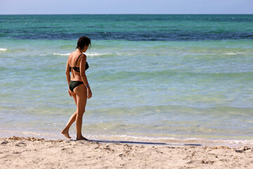 Slim tanned woman in bikini walking by the sand on sea beach. Vacation on tourist resort