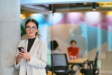 In a modern glass office, a contemporary businesswoman effortlessly multitasks, using her smartphone, while surrounded by colleagues, illustrating the dynamics of corporate life and technology