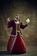 Gray-haired smiling man, medieval royal person, king in female dress eating instant noodles against vintage green background. Concept of historical retrospectives, fashion, provoking projects, food