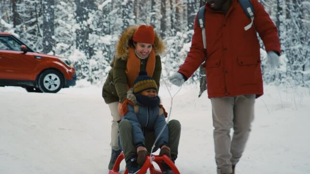 Long shot of multi-ethnic mother and father riding their son on sled in snowy winter with red car in background