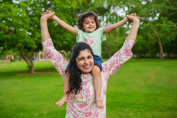 Happy young indian mother carrying child daughter on her shoulders at summer park.Family and motherhood concept.