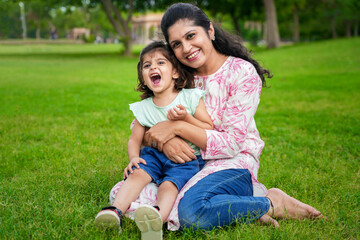 Happy playful young indian mother and  little daughter sitting at summer park having fun together....