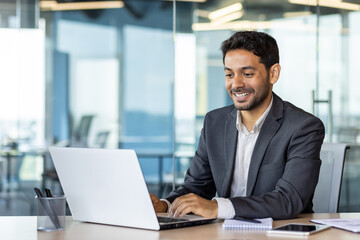 Fototapeta na wymiar Portrait of young arab businessman inside office, man typing on laptop smiling, boss happy with results of financial success at workplace.