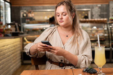 Charming blonde woman with beautiful smile reading good news on mobile phone during rest in coffee shop
