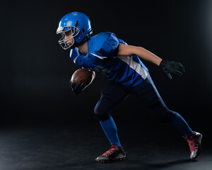 Full length portrait of a man in a blue american football uniform against a black background. Sportsman in a helmet with a ball. 