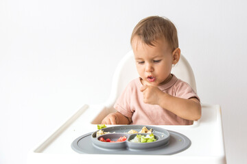 Cute little baby sits in a high chair on a white background and eats complementary foods with interest, concept of baby feeding and self-feeding blw