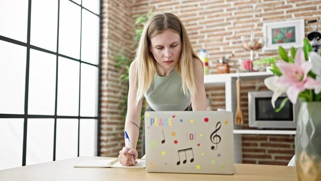 Young blonde woman using laptop writing on notebook studying at dinning room