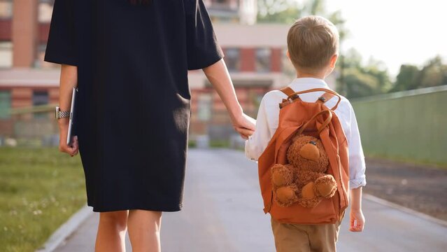 Back view mother leads her son by hand to school. Family is holding hands. Baby boy in school uniform. White shirt and backpack. Preschool child goes to first grade. September. New academic year.