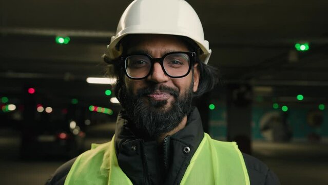 Portrait Indian man handyman worker contractor mechanic guy in helmet and glasses smiling indoors parking factory warehouse manufacture. Close up Arabian male builder engineer fitter in hard hat smile