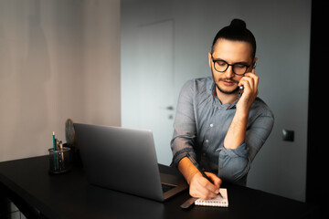 Portrait of young serious businessman with hair bun, working home at laptop, makes notes in notepad, talking on smartphone with clients, wearing eyeglasses and shirt. Background of grey wall.