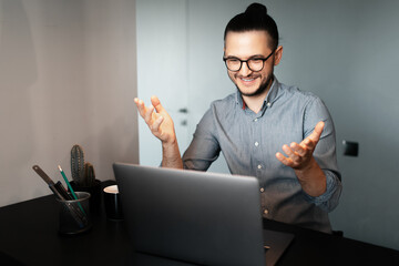 Portrait of young happy businessman working home at laptop, feeling satisfied about work, talking with people through webcam conference, wearing eyeglasses and shirt on background of grey walls.