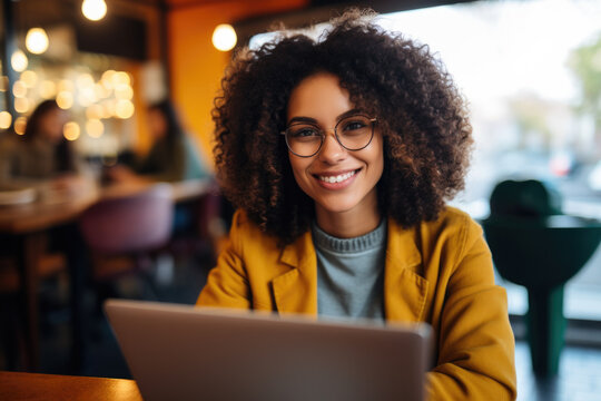 Beautiful young woman with curly hair wearing yellow jacket sitting in cafe working with laptop.