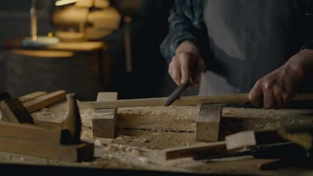 A woodworker works with a hand tool to process a piece of solid wood, a woodworker in special clothing works in a workshop using hand tools to process wood. Handmade crafts of wooden objects