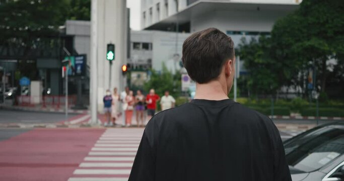 Male pedestrian stands at pedestrian crossing of road, waiting green light at traffic light allowing him to cross road. Rear view of man crossing the road at pedestrian crossing with a traffic light
