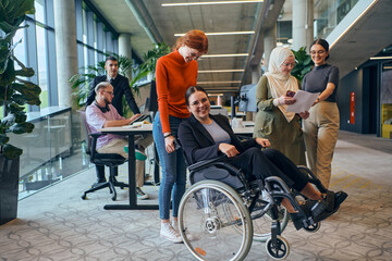 A diverse group of business colleagues is having fun with their wheelchair-using colleague,...