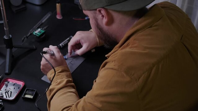 A young man in a mustard shirt and cap works in dim light at a table and solders wires with a soldering iron. Working with a soldering iron from which smoke comes, engineering activities.