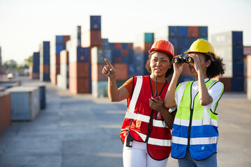 young female African factory workers or engineer using binoculars and pointing to something in containers warehouse storage