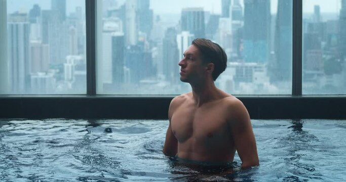 Caucasian man relax in the roof top swimming pool in luxury hotel in big city. Young man enjoying in luxury swimming pool on the roof of skyscraper on background of city. Summer vacation concept