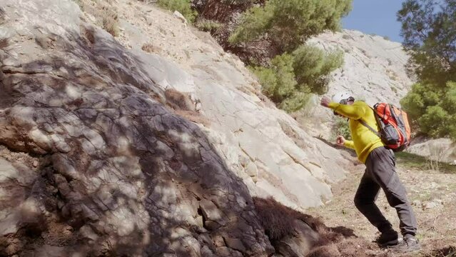 tourist in yellow jacket with orange backpack climbs cliff, mature man in safety helmet, rock climber, extreme people lifestyle, mountaineering, life insurance, difficulties in hike, extreme sports