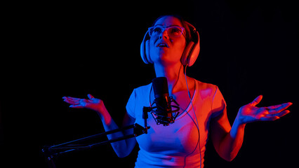 Caucasian woman in glasses and headphones sings into a microphone in neon light on a black background. An emotional girl is recording a song in a recording studio