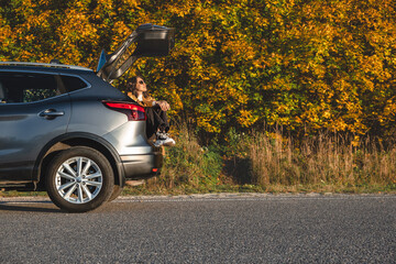 a dreamy woman sits in the trunk of her car and enjoys the warmth near the autumn forest