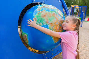 A little girl of 7-8 years old with long hair, dressed in a pink T-shirt and shorts, smiles and hugs the globe - the globe in an amusement park.