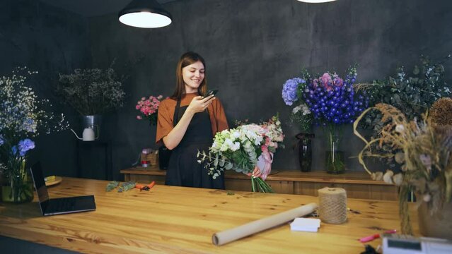 Happy smiling woman florist photographs a bouquet with her phone, the result of her work. Floristry and retail flower trade