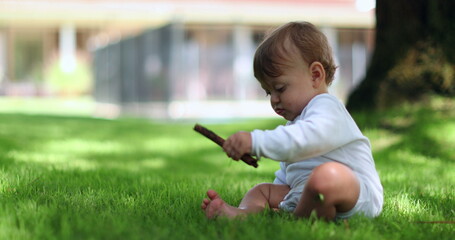 Adorable baby sitting outside in home lawn holding stick. Cute infant toddler playing with stick outdoors - obrazy, fototapety, plakaty