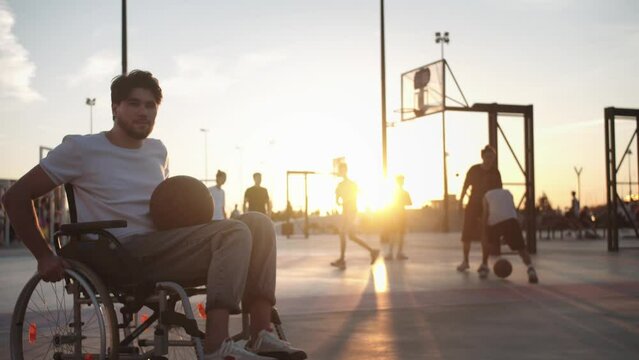 A man in a wheelchair rides in the frame with a basketball court and players in the background